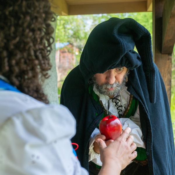 An actor portraying the wicked stepmother, wearing a cowl, hands an apple to Snow White.