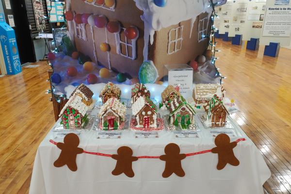 Gingerbread houses displayed on a table with gingerbread men garland, and a very large gingerbread house behind the table. 