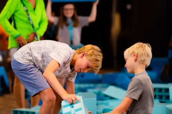 In the background, a woman in a bright green jacket looks over the piles of blue mega bricks. A teenage girl is also in the background holding a blue mega brick over her head. In the foreground, two young boys are putting together two blue mega bricks.