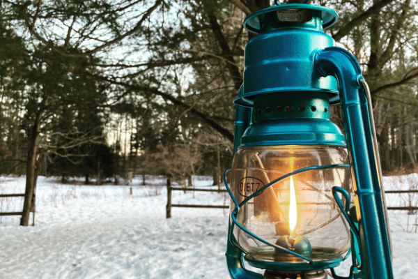 Teal lantern in the foreground, it is lit. It stands in the snow. In the background are pines trees, snow, and an open wooden fence.