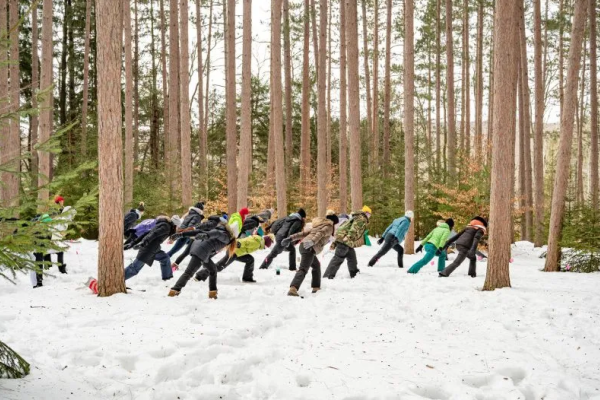 People dressed in winter gear doing a yoga pose with snow and pine trees in the background.