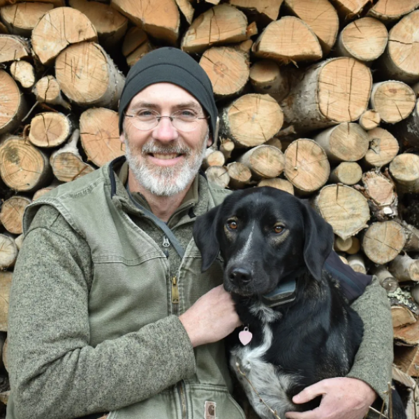 Presenter Dave Hall sits in front of a huge wood pile with his arm around his dog. His dog is a short hair with black fur and a white chest.