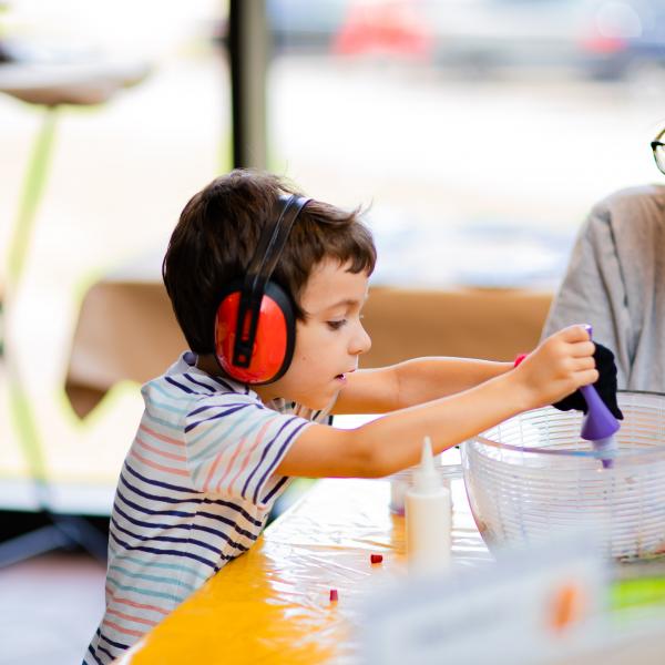 Small child sitting at craft table squeezing a purple paint bottle into a salad spinner while wearing red headphones. A young woman, sitting at the end of the table, watches the craft making.