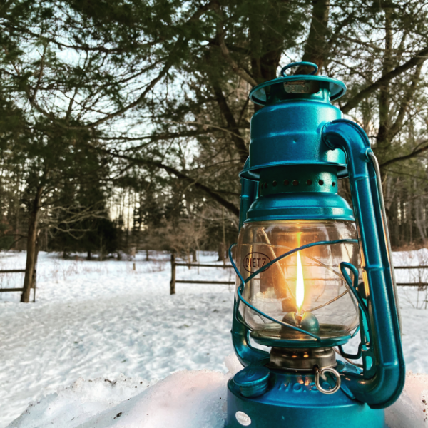 Teal lantern in the foreground, it is lit. It stands in the snow. In the background are pines trees, snow, and an open wooden fence.