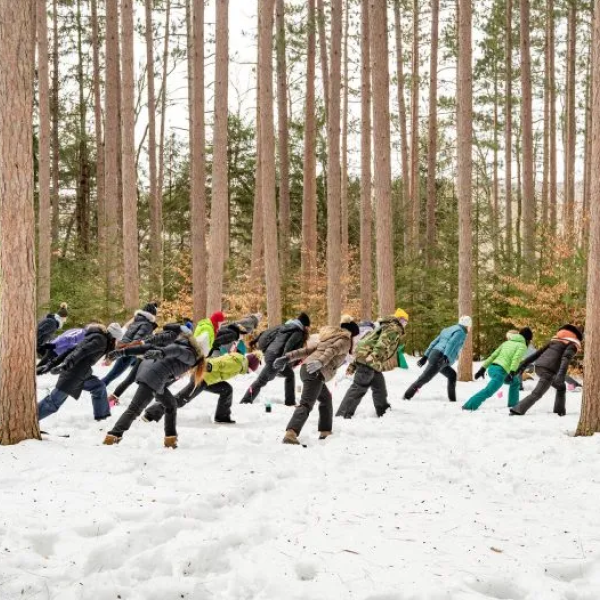 People dressed in winter gear doing a yoga pose with snow and pine trees in the background.