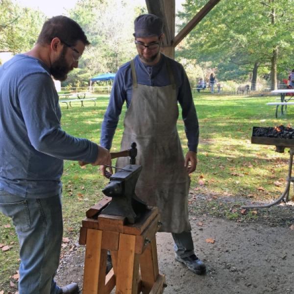 First man with a blue shirt, a beard, and safety goggles is hammering a piece of metal. The second man in a blue shirt, a beard, and safety goggles is watching the first man. Second man is the teacher and they are both outside under a pavilion.
