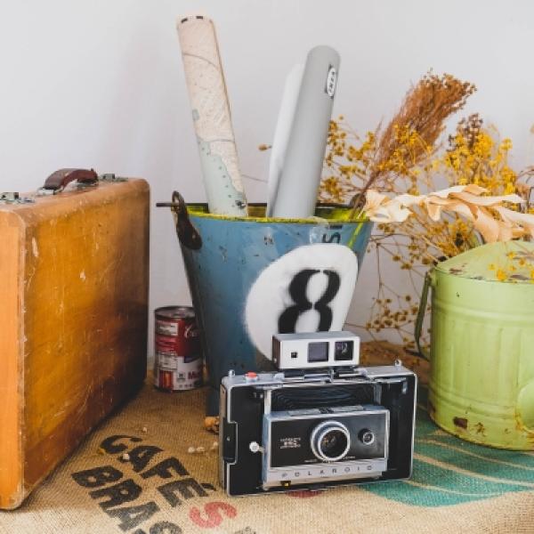 A photograph of a suitcase, an old camera, a metal bucket holding rolled up paper, and a watering can. There is a can of Campbell's soup behind and between the suitcase and the metal bucket.