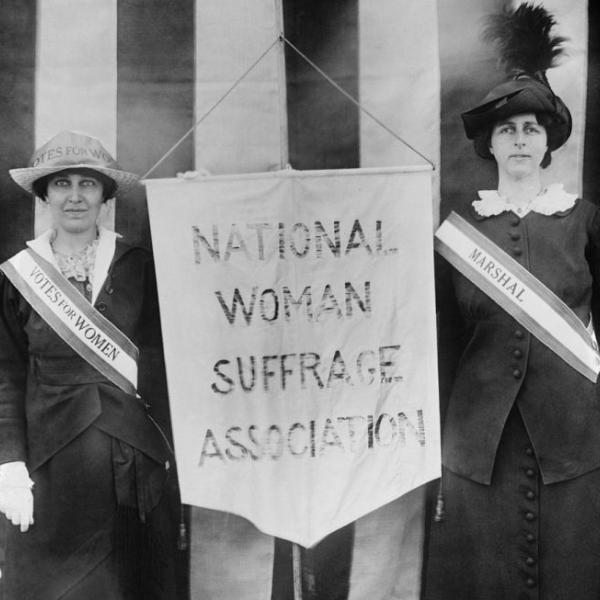 Black and white photograph of two women standing with a banner in between them that says "National Woman Suffrage Association".